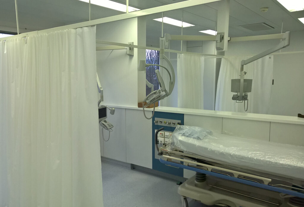 Ein steriler Operationssaal mit einem Rollbett, einer Telefonkonsole und Computerbildschirmen an schwenkbaren Armen sowie weissen Vorhängen an der Schiene.